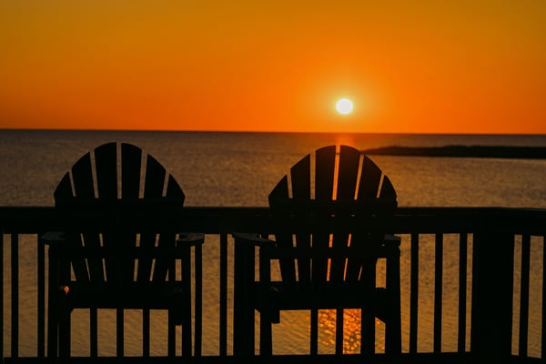 Two Adirondack chairs silhouetted against a vivid orange Gulf sunset over calm water