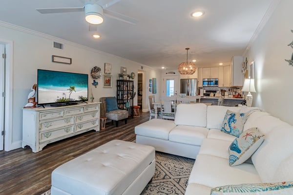 Open-plan living room and kitchen with cream sofa, white ottoman, TV on dresser, and stainless steel appliances