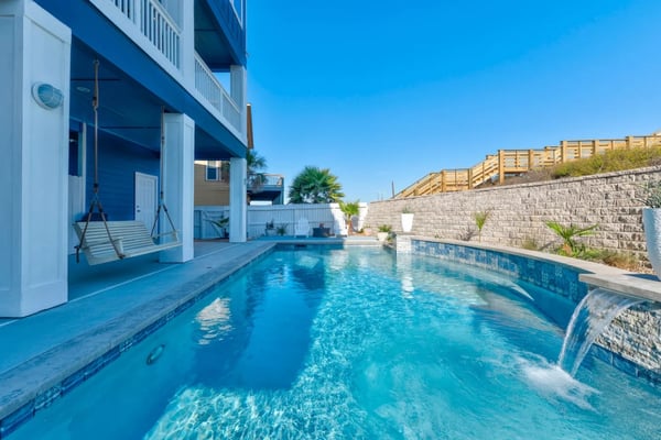 Backyard swimming pool with waterfall feature, swing, and blue beach house under clear sky