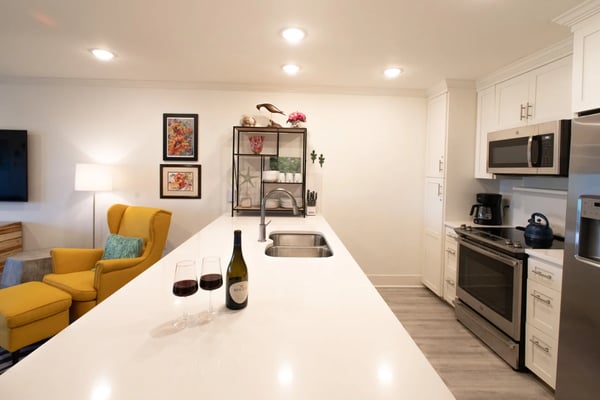 Modern kitchen and living area with white cabinetry, stainless steel appliances, long white island with wine bottle and glasses, yellow armchair, and open